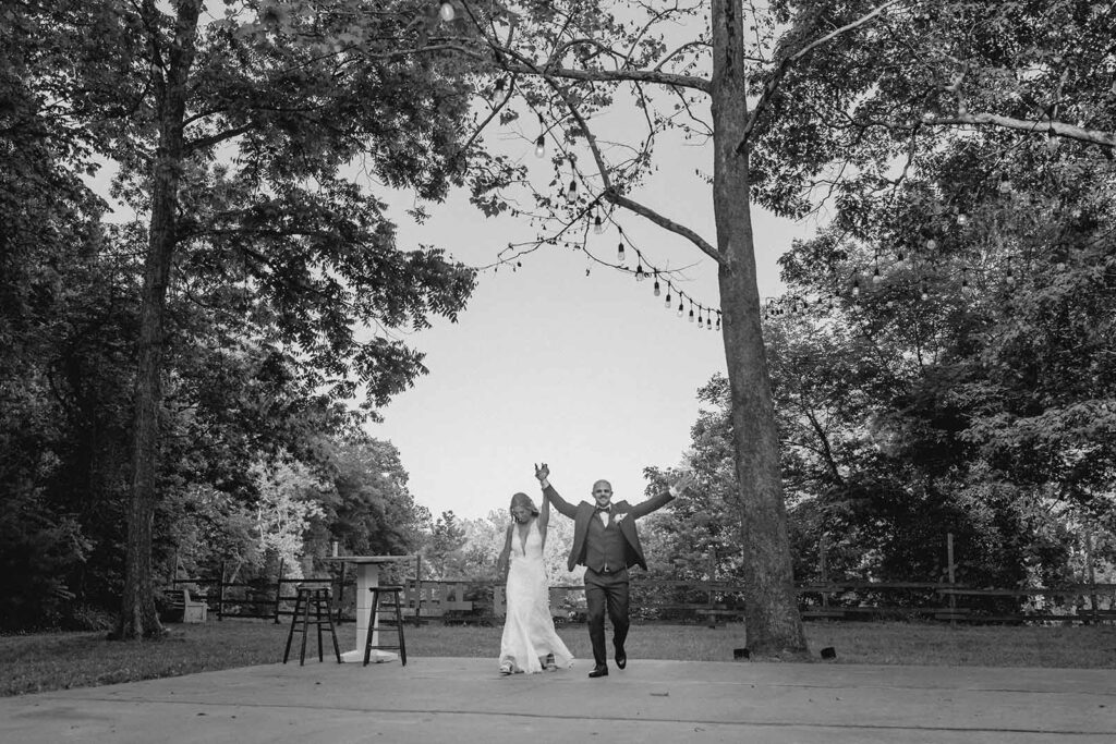 Wide shot of bride and groom holding hands to the air for grand entrance under canopy of tall trees