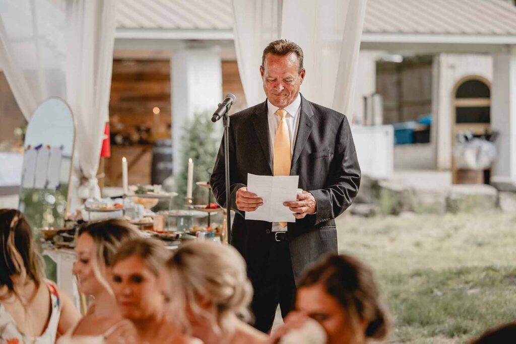 Father of the groom in black suit and yellow tie reads speech during summer evening reception
