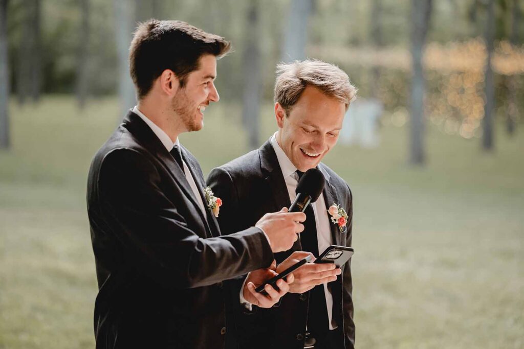 Co-best men in black suits laugh into the microphone while giving wedding speech together