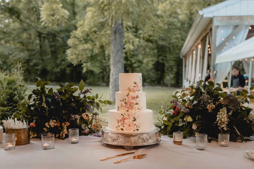 Three tier white wedding cake with wildflower icing details sit between two floral arrangements on white dessert table