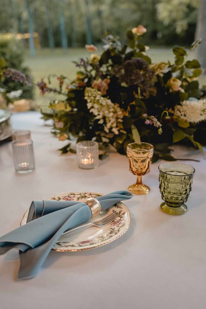 Desert table close up of antique painted place, blue napkin and vintage colored glasses on white tablecloth in front of an arrangement of wildflowers
