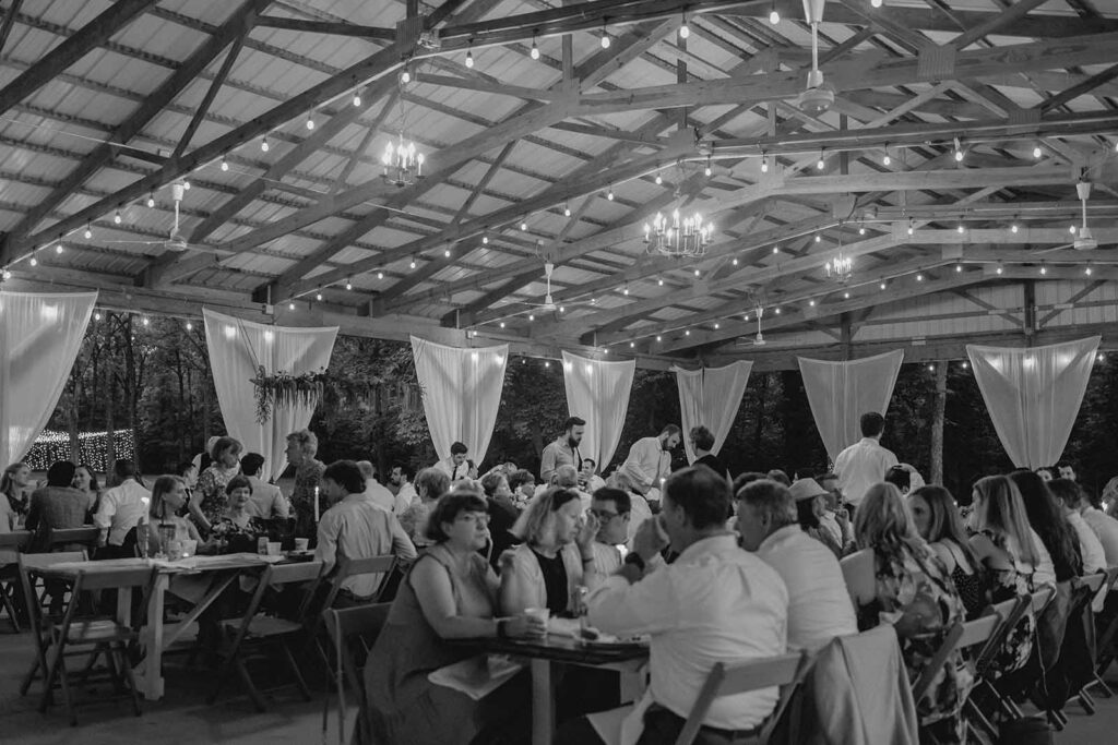Black and white wide shot of intimate wedding dinner under a pavilion of lined with string lights