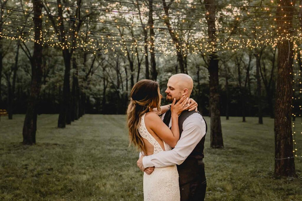 Bride and groom embrace under a canopy of string lights in a grove of trees during blue hour
