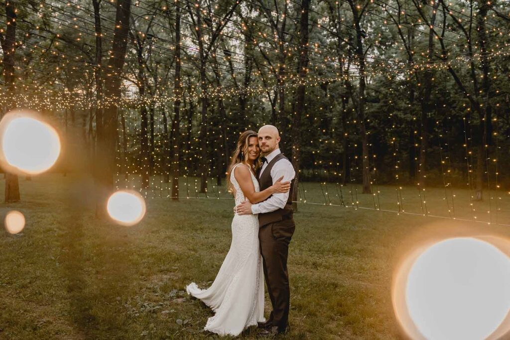 Bride and groom look toward the camera surrounded by close and far string lights in a grove of trees during blue hour