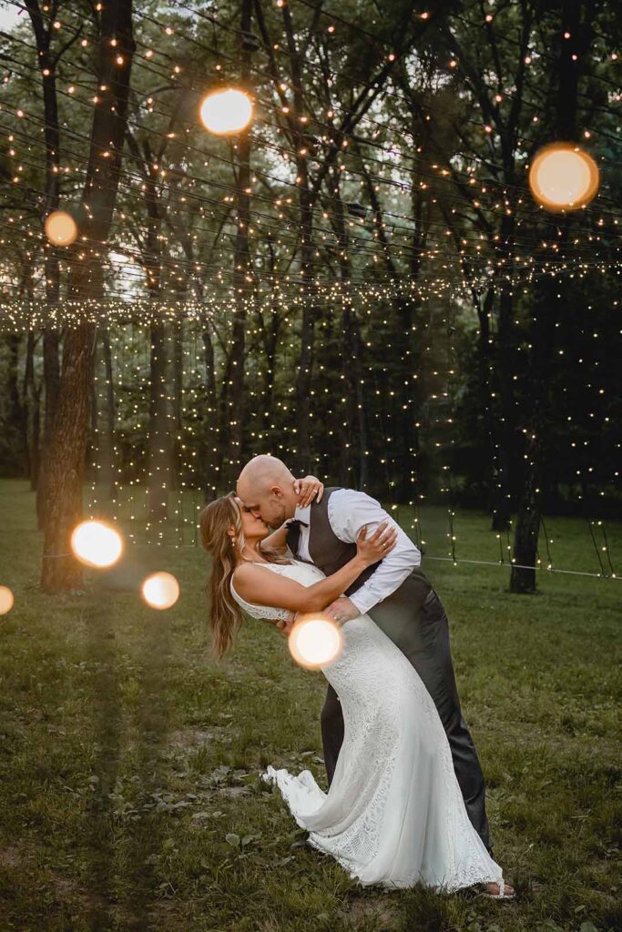 Bride and groom dip back into a kiss against a background of string lights in a grove of trees during blue hour