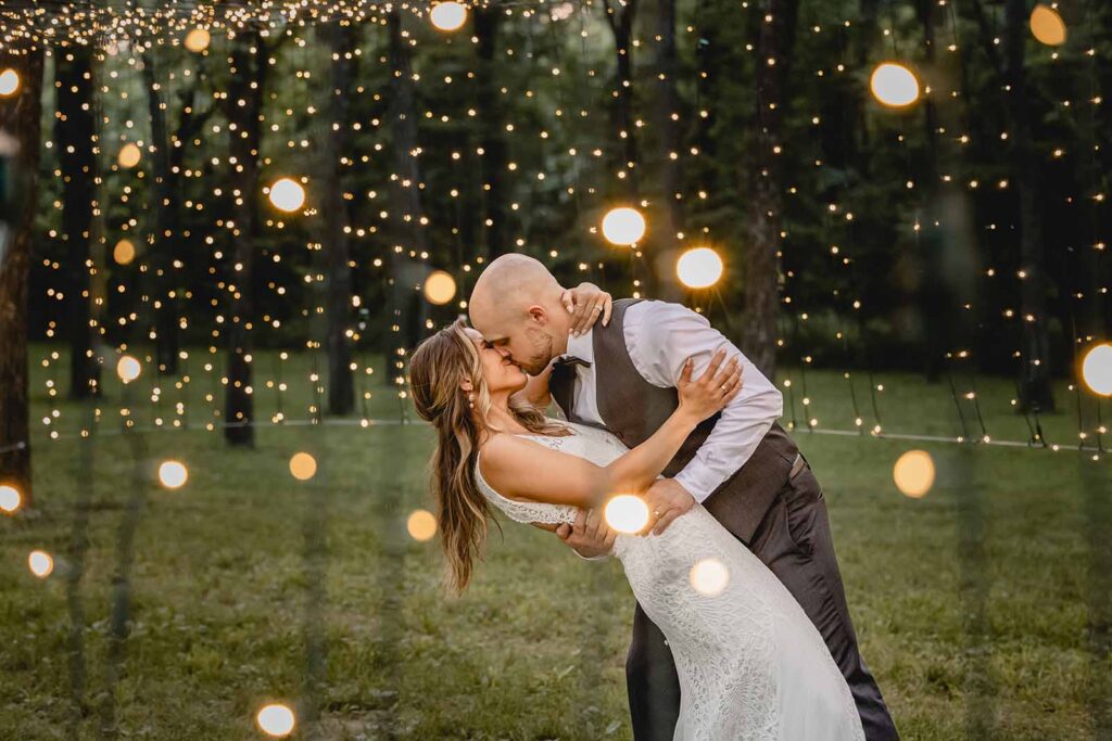 Bride and groom dip back into a kiss against a background of string lights in a grove of trees during blue hour