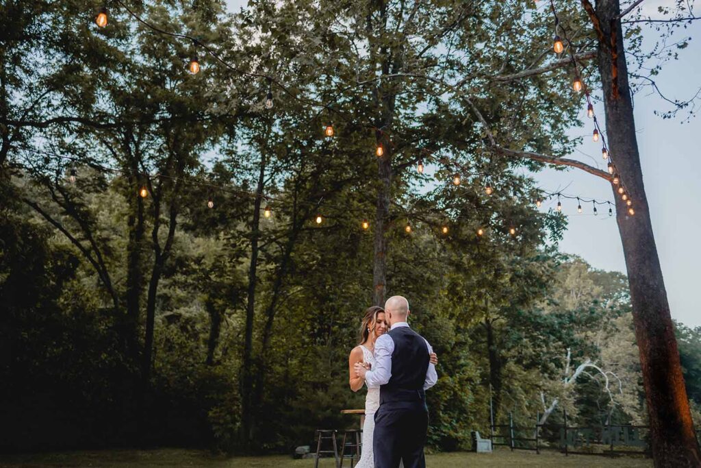 Wide shot from below of bride and groom swaying close during first dance outside during blue hour under string lights and trees