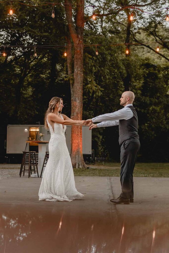 Bride and groom hold hands outstretched during first dance outside during blue hour with string lights twinkling above