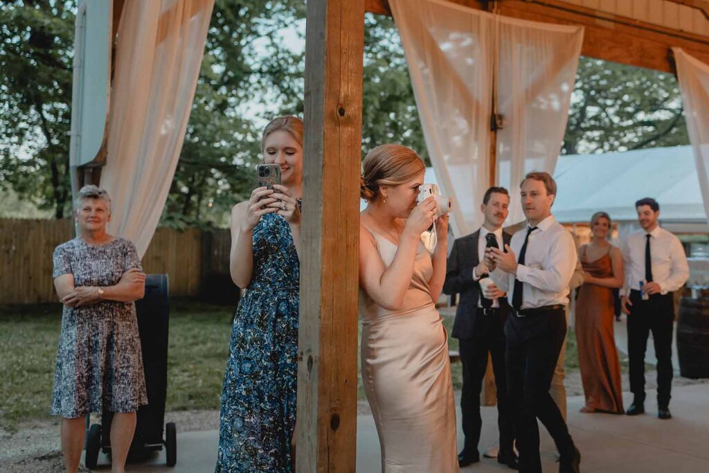 Candid shot of two wedding guests leaning against pavilion pole, one taking photos of the dance floor the other of guests