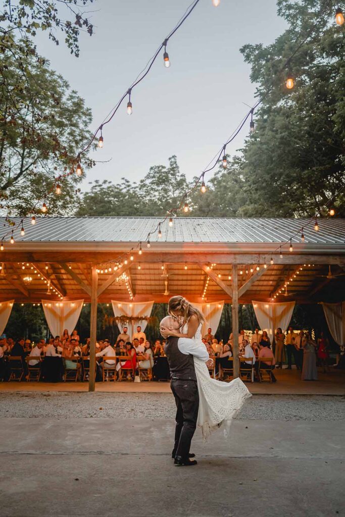 Groom holding bride up in a lift kiss under a sea of string lights with their pavilion of guests in the background