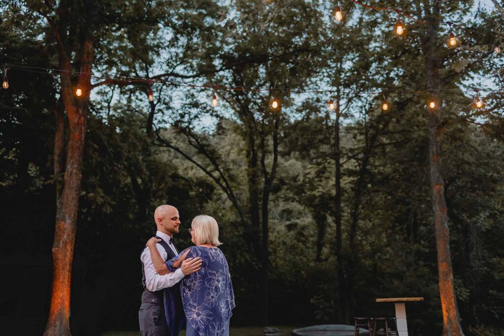Wide shot from below of groom and his mother during first dance outside during blue hour under string lights and trees