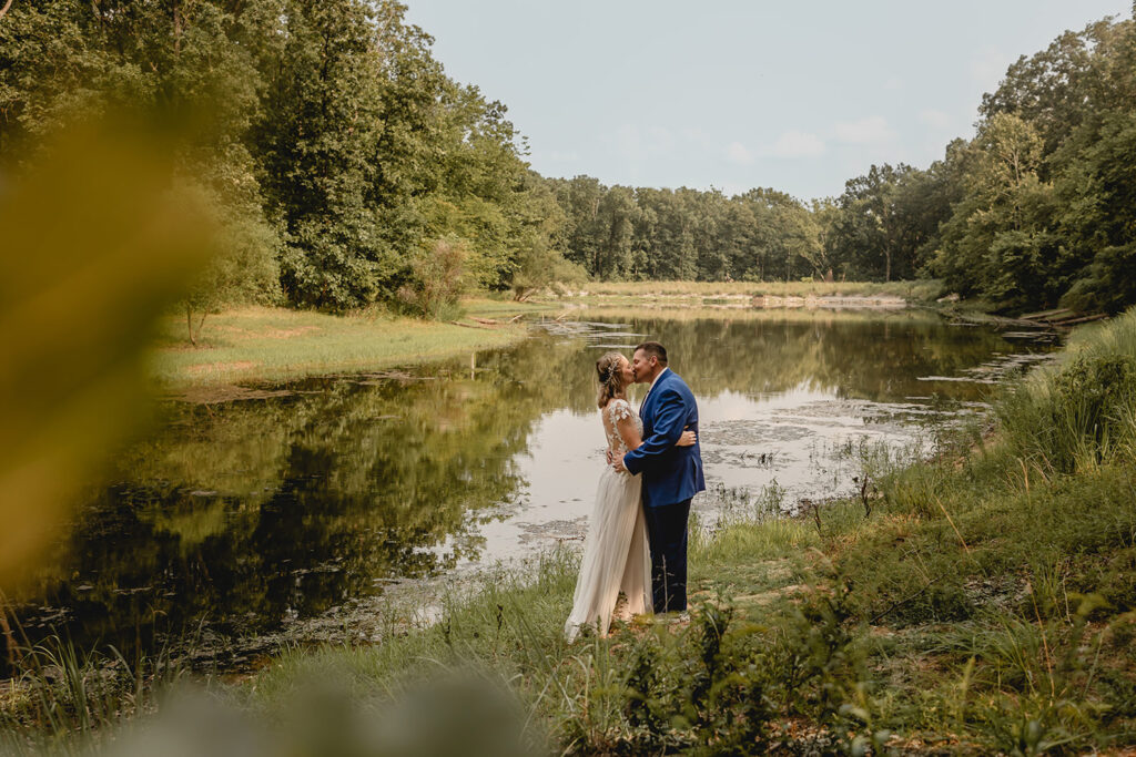 Wide shot between green foliage of  bride and groom kissing in front a small lake at Quail Ridge Park
