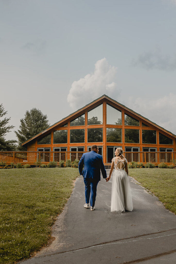 Newlyweds hold hands and walk away from the camera towards their glass lodge venue