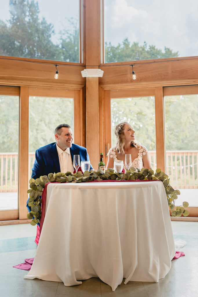 Newlyweds smile at their small sweetheart table in front of a wall of windows