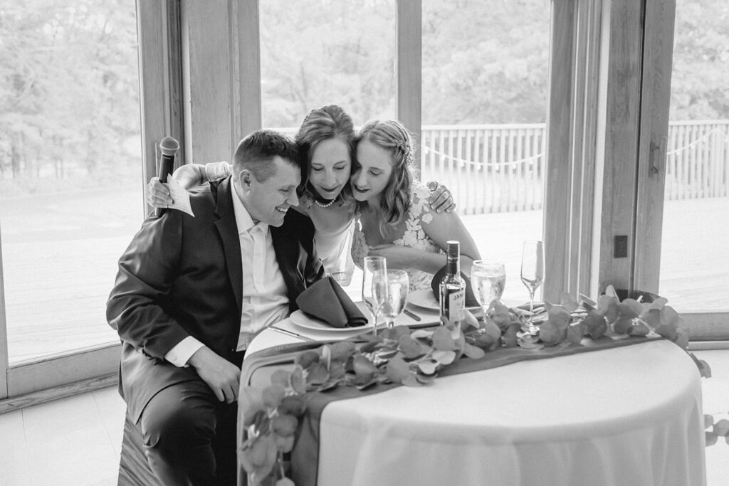 black and white shot of bride's mother hugging the newlyweds at their sweetheart table