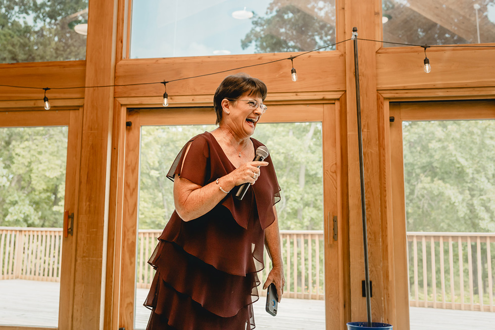 mother of the groom laughs with a microphone in hand as she delivers her wedding speech