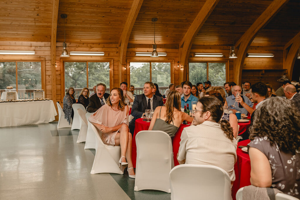 wedding guests listen to toasts while seated around their tables in wooden lodge