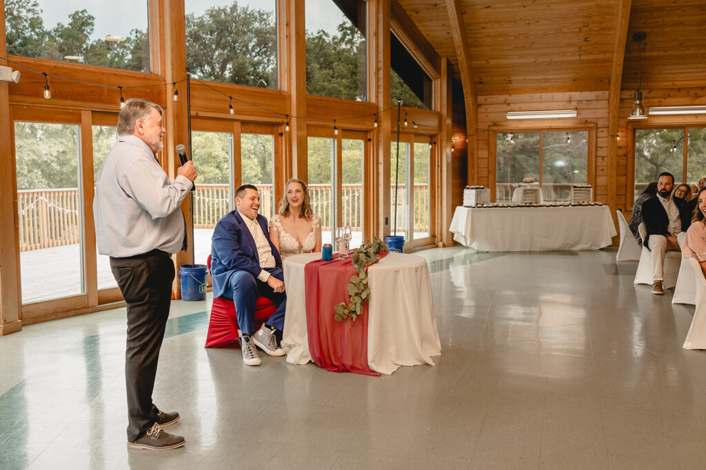 Wide shot of lodge room as groom laughs at the sweetheart table while his father gives a toast