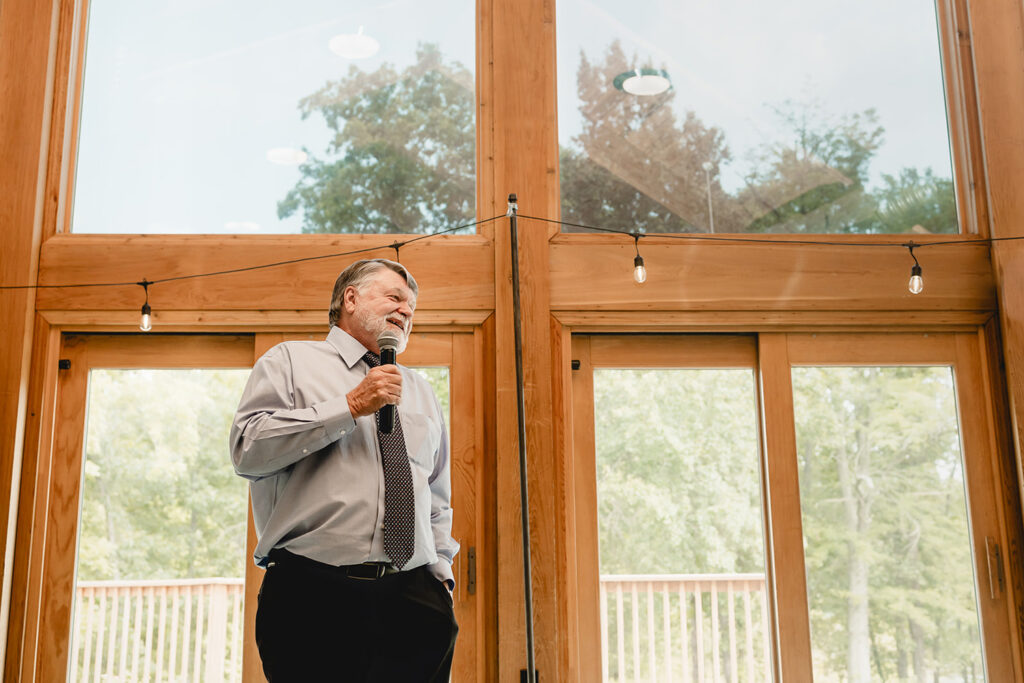 Father of the groom giving a wedding toast in front of a wall of window to outside greenery