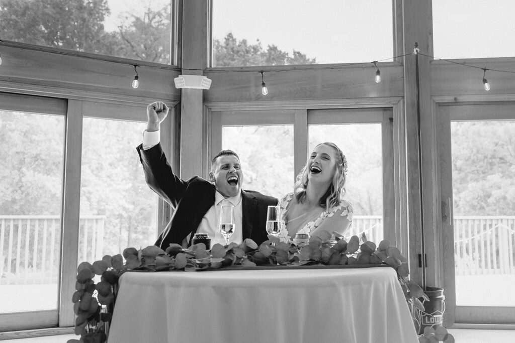 black and white shot of groom pumping his fist in excitement as he and his wife life in reaction to wedding speech