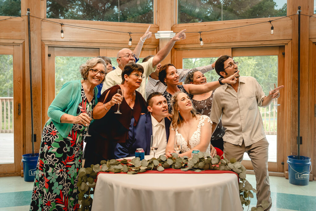 bride and groom seated at their sweetheart table while a group of guests behind point and look shocked at something in the distance 