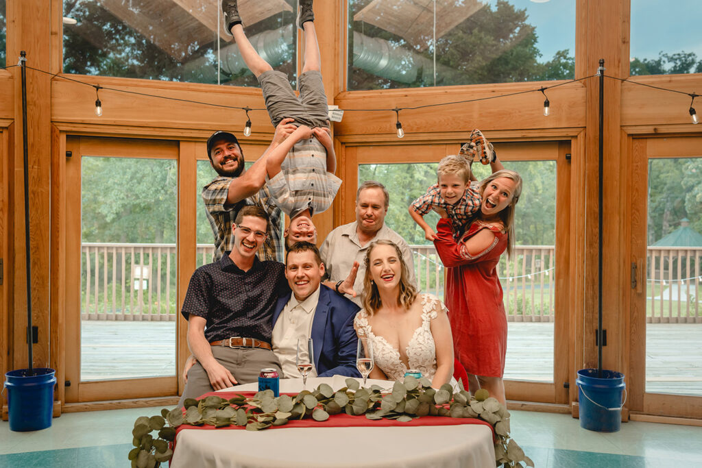 bride and groom seated at their sweetheart table while a group of guests behind hold their children upside down in the air