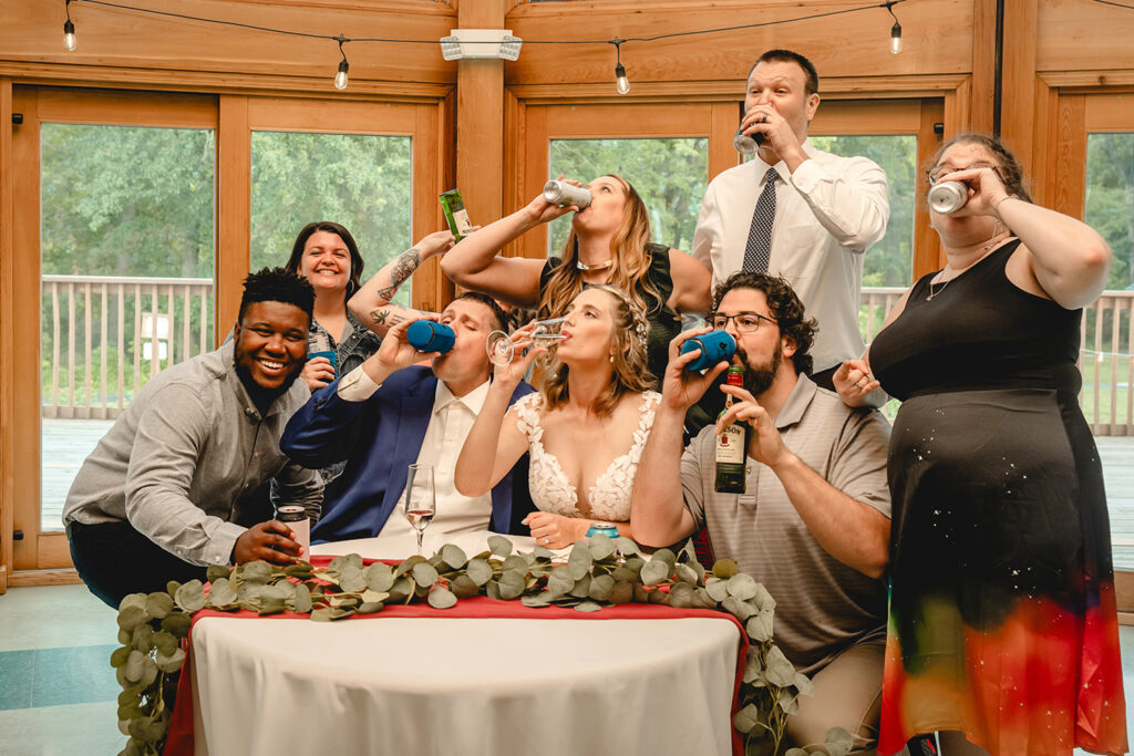 bride and groom seated at their sweetheart table while a group of guests behind chug their drinks
