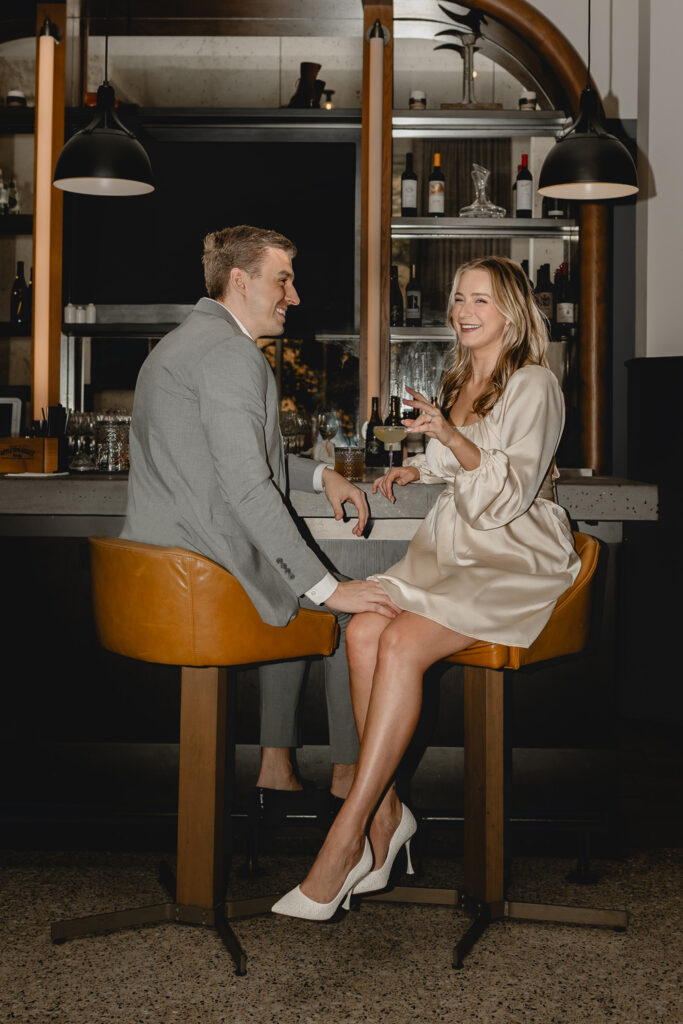 young man in gray suit puts his hand on his girlfriends leg and looks to her as she smiles towards the camera from The Last Hotel STL  lounge bar stools
