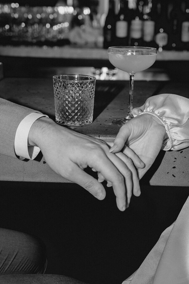 two hands rest in each other draped along the edge of a bar in front of cocktail glasses