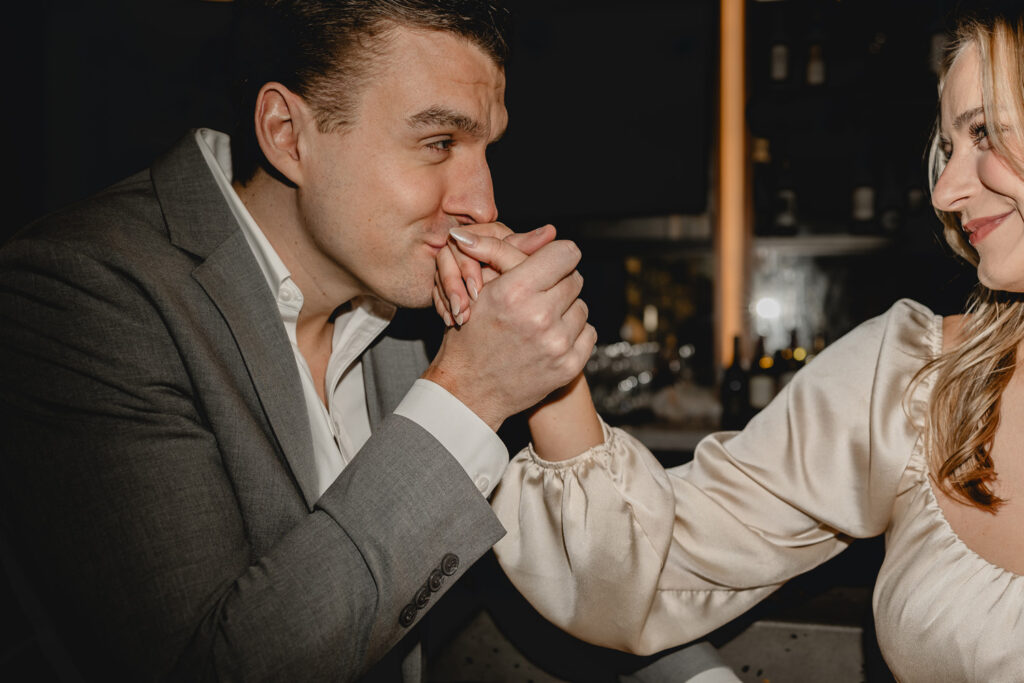 a young man in a gray suit and white dress shirt kisses his girlfriend's hand as she smirks from the side of frame