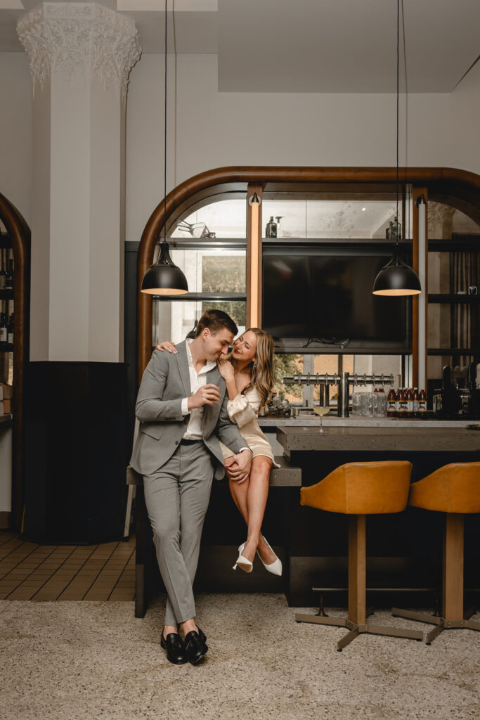 young man in a gray suit leans against the hotel bar his girlfriend sits upon and hugs him from behind 