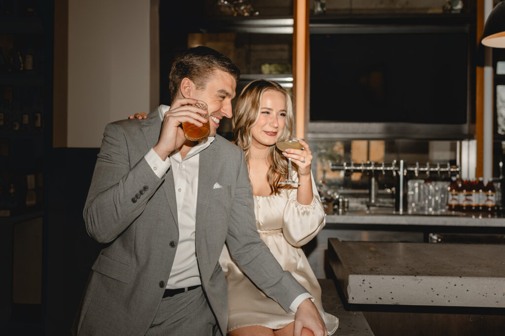 young dressy man holds a cocktail glass as he laughs towards his girlfriend holding her drink by his side