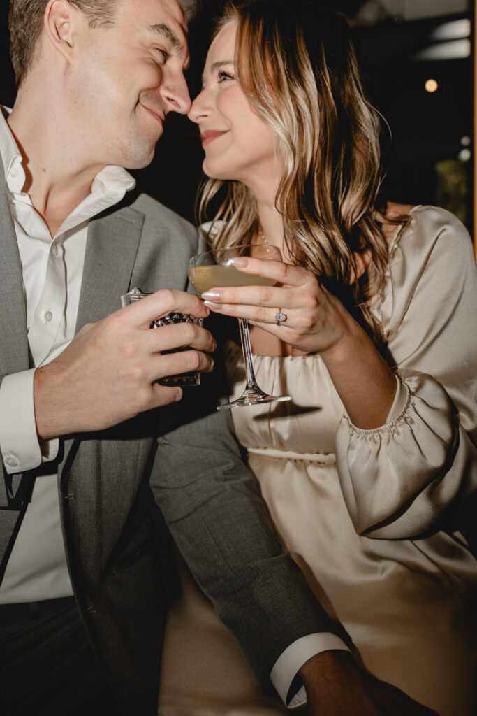 dramatic direct-flash shot of a young couple nuzzling  noses and clinking cocktails glasses in the foreground to feature the girlfriend's engagement ring 