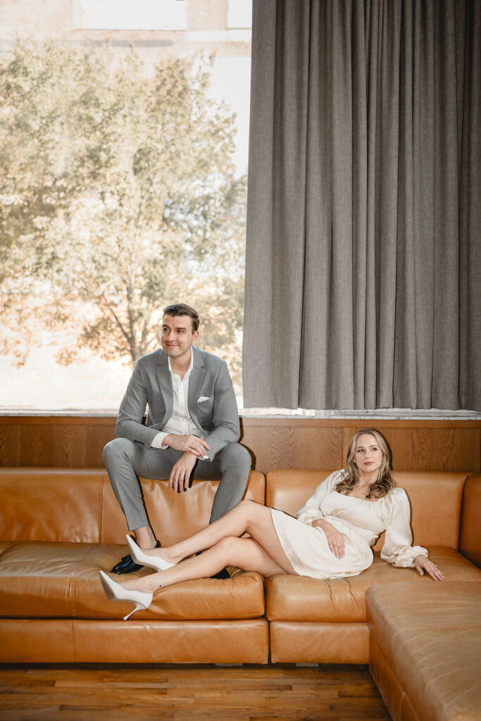 young stylish man in a suit sits on the top edge of retro leather couch as woman in a white dress and heels leans by his feet
