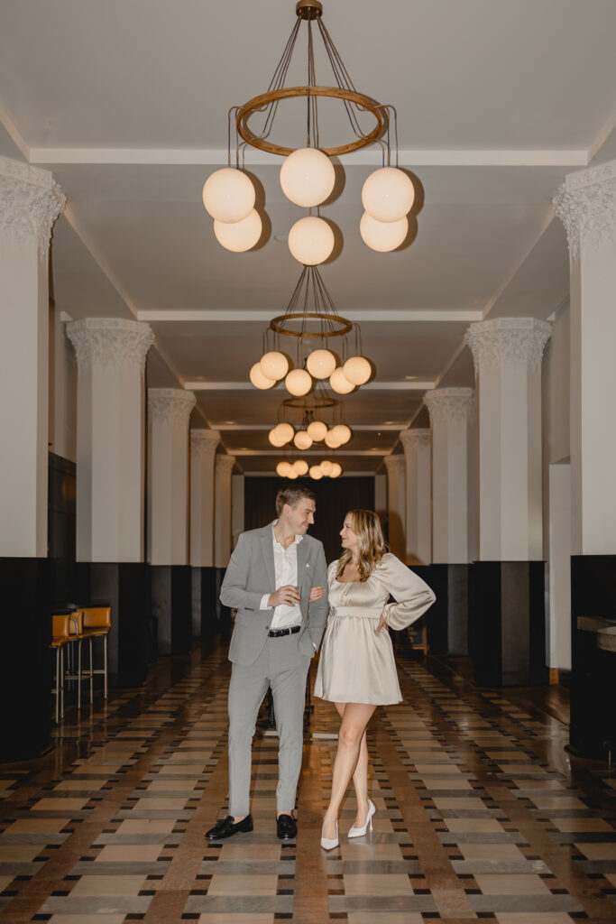 couple standing centered in a stylish hotel lobby hallway lined with columns and orb chandeliers
