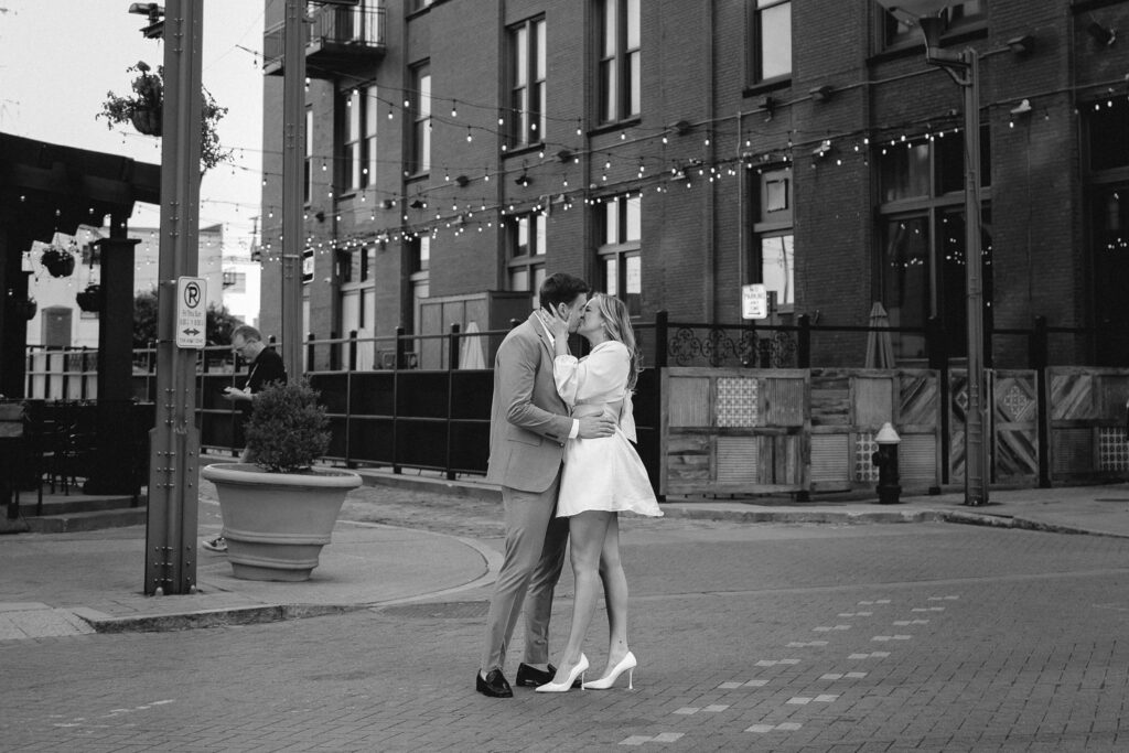 stylish couple dressed in suit and heels kid in the middle of a downtown street with a brick building and string light over a patio behind