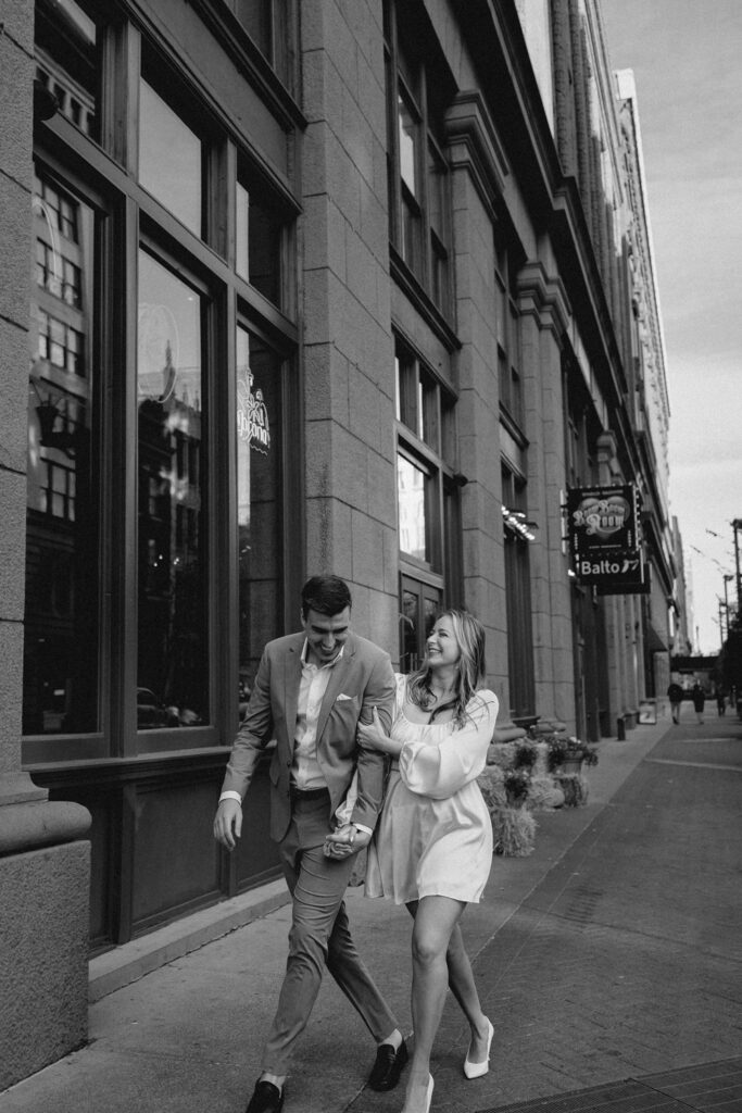 black and white shot of young stylish couple holding hands, and giggling mid-walk on a downtown street