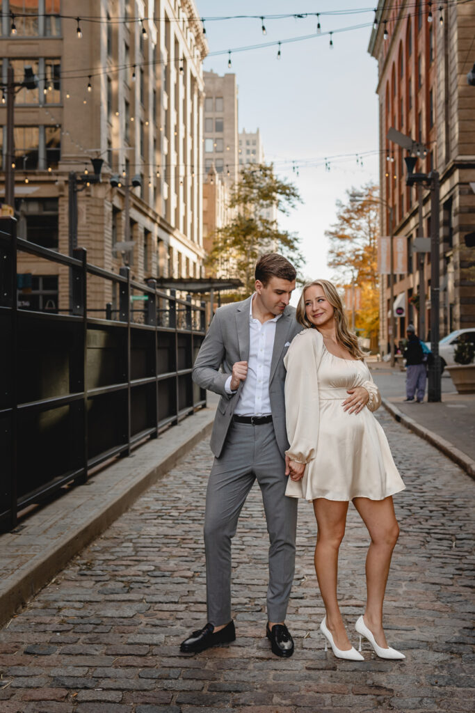 young couple centered in a cobblestone alleyway looking towards each other and accentuating their outfits surrounded by tall tan buildings