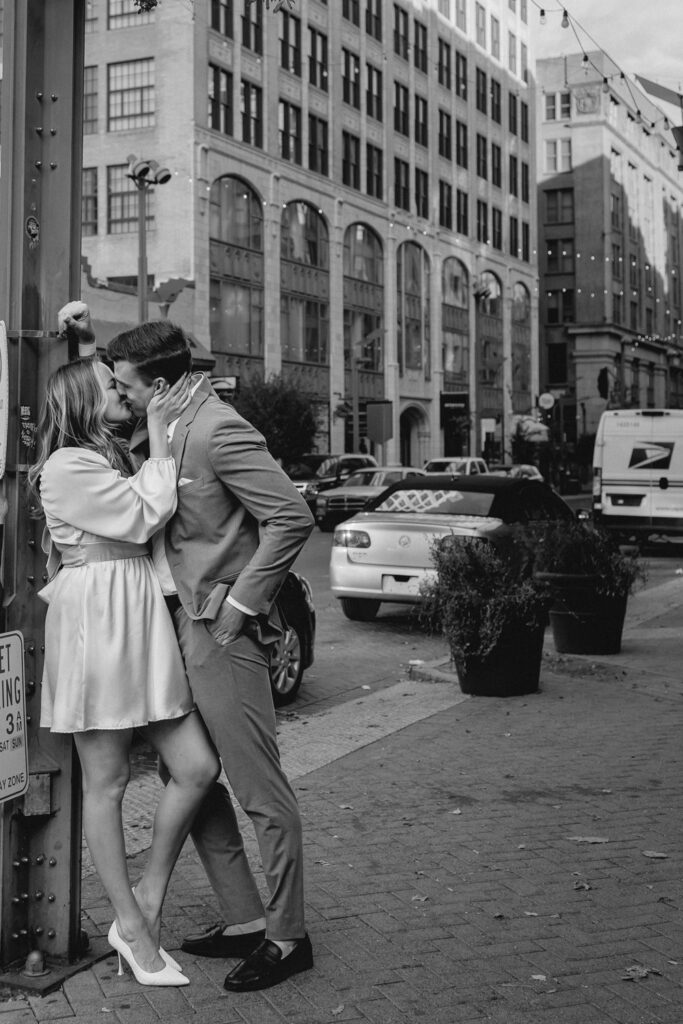 a young couple lean against a light pull and kiss in a moody black and white portrait that feels like a scene from New York City