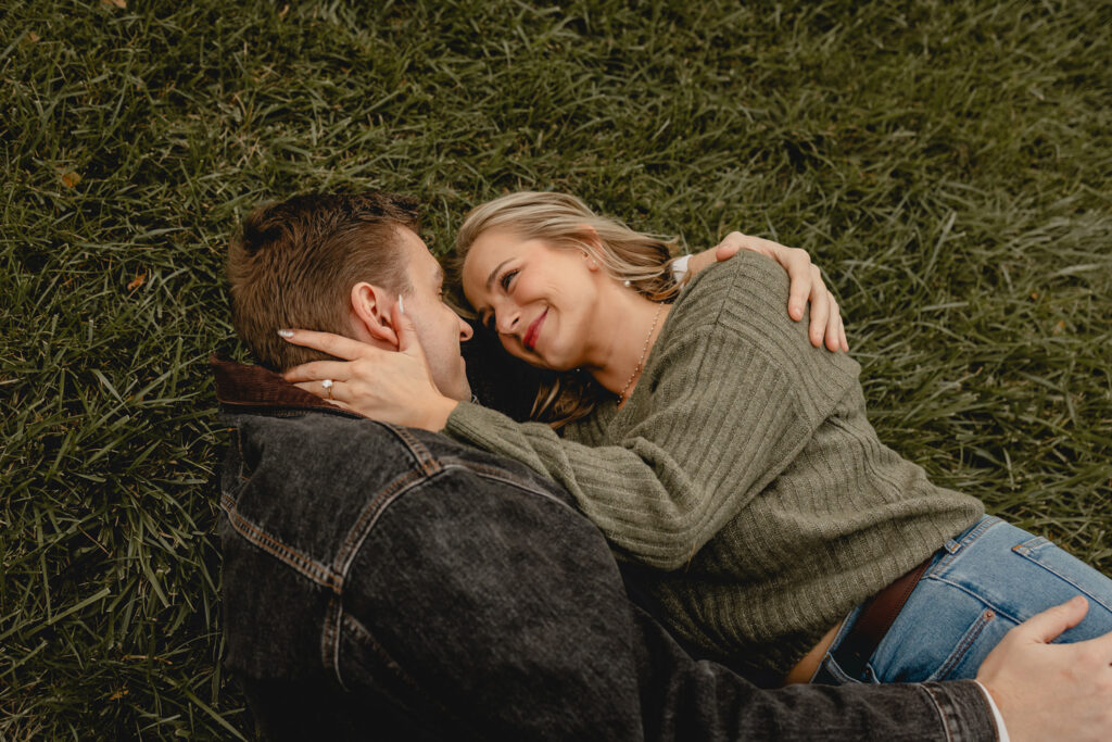 young man in white tshirt and dark denim jacket and woman in chunky green sweater cuddle in lush grass in Forest Park