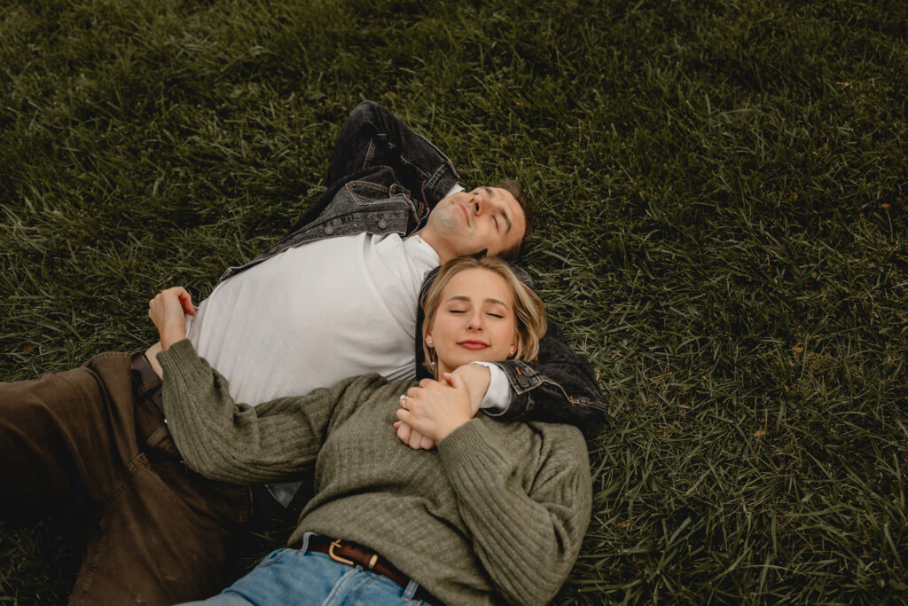 young man in white tshirt and dark denim jacket and woman in chunky green sweater with jeans lay on their backs in the grass with eyes closed as if daydreaming