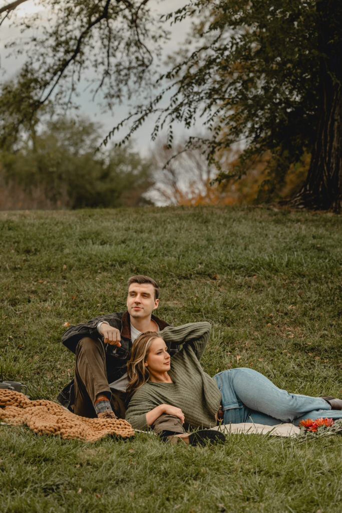 women laying on her side brushing back her hair as her boyfriend's sits up behind looking off the opposit direction on a grassy hill