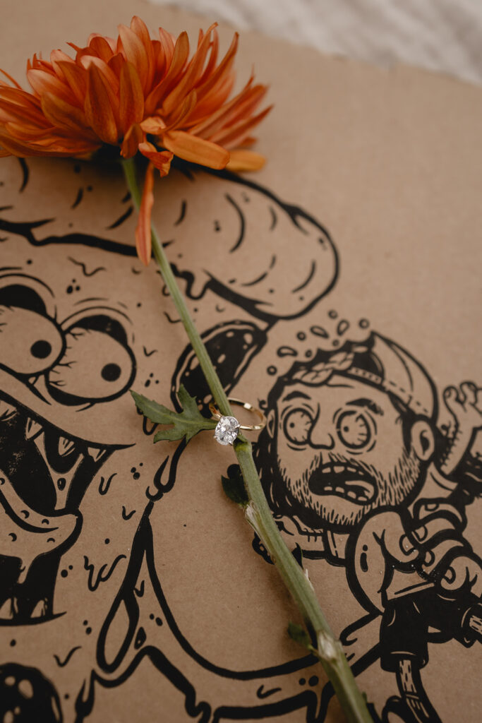 the long stem of an orange wildflower sits inside of gold oval diamond engagement ring untop of funny pizza box top illustration