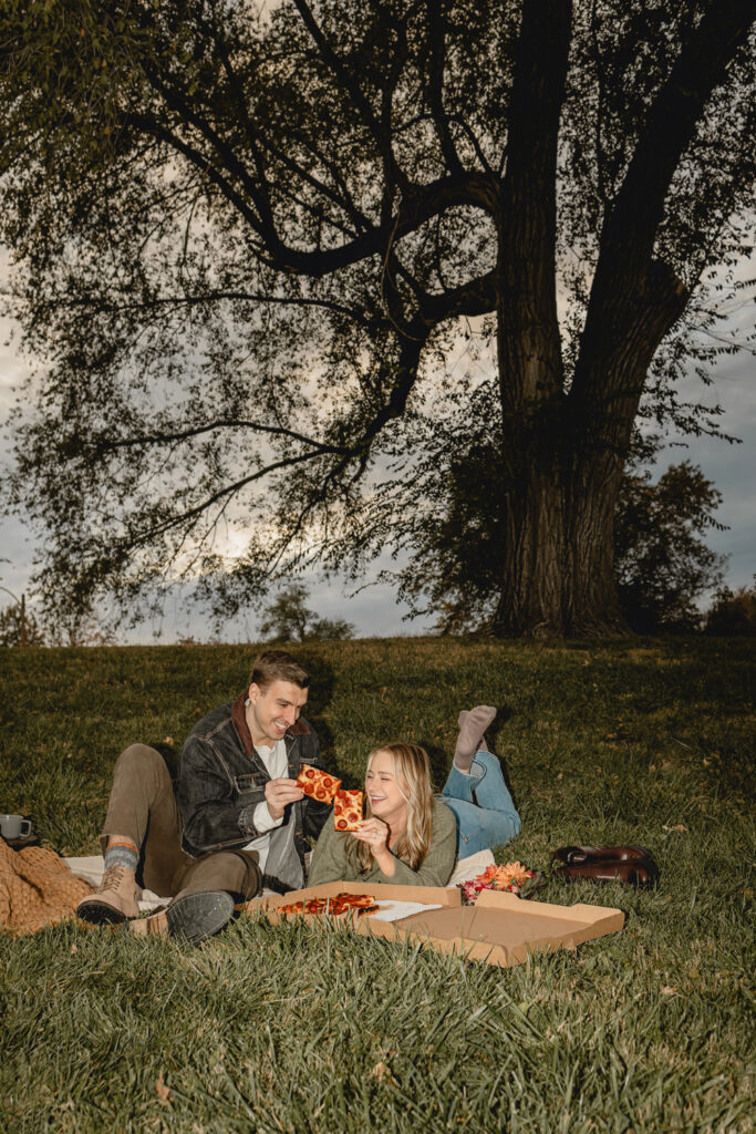 direct flash shot of seated man and woman laying on her stomach with her feet up cheers-ing two slices of pizza on a sunset grassy hill