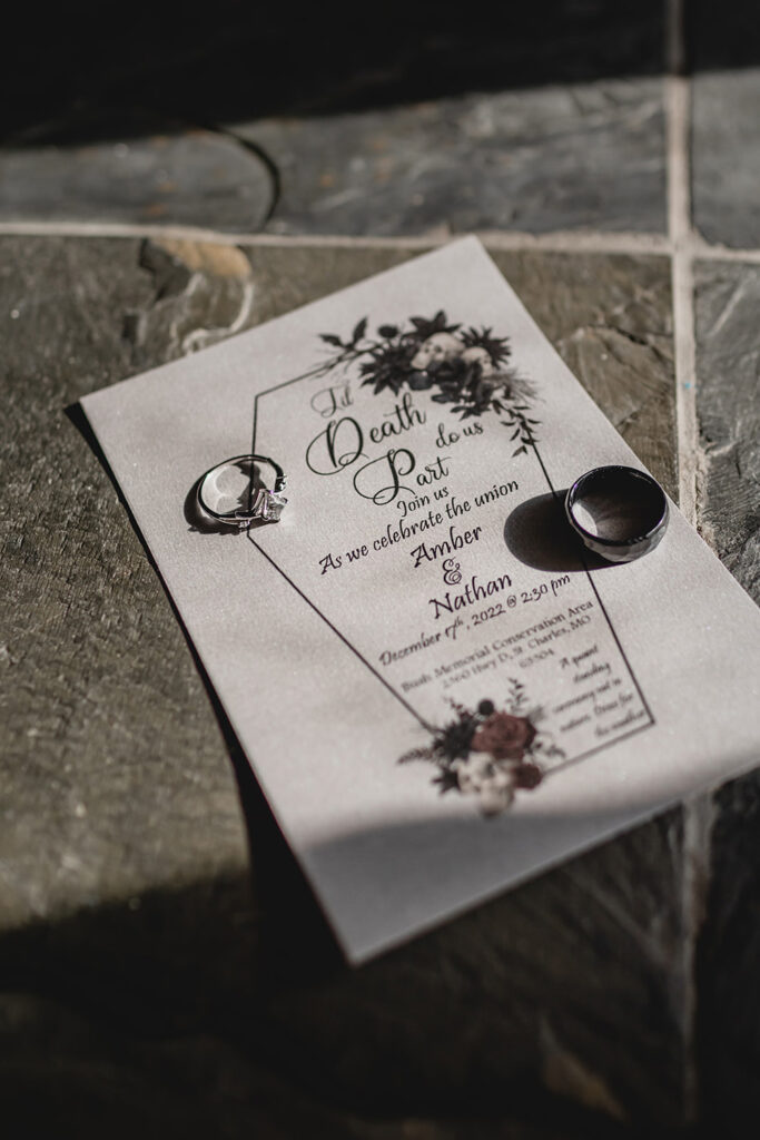 two wedding rings sit atop a metallic silver wedding invitation that says "Til Death Do Us Part" inside of a coffin shape