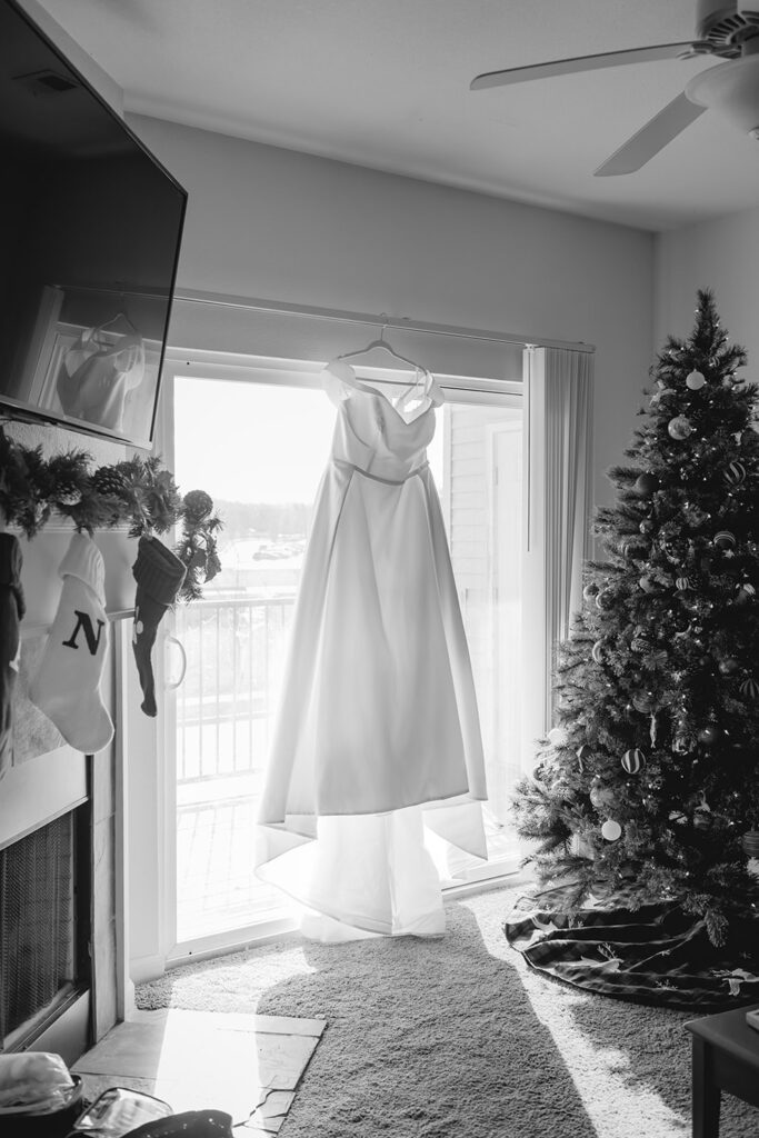 a simple white wedding dress hangs on the curtain rod above a large sliding glass door in between a fireplace with stockings and christmas tree