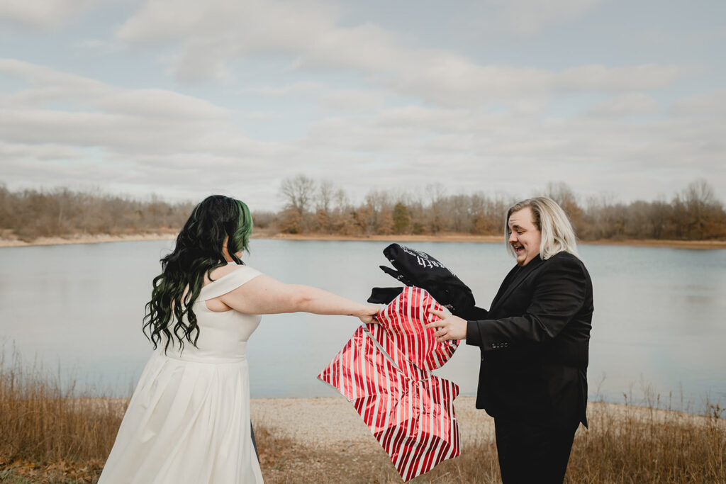 bride reaches to grab striped Christmas wrapping paper as the groom unwraps her gift in front of a lake