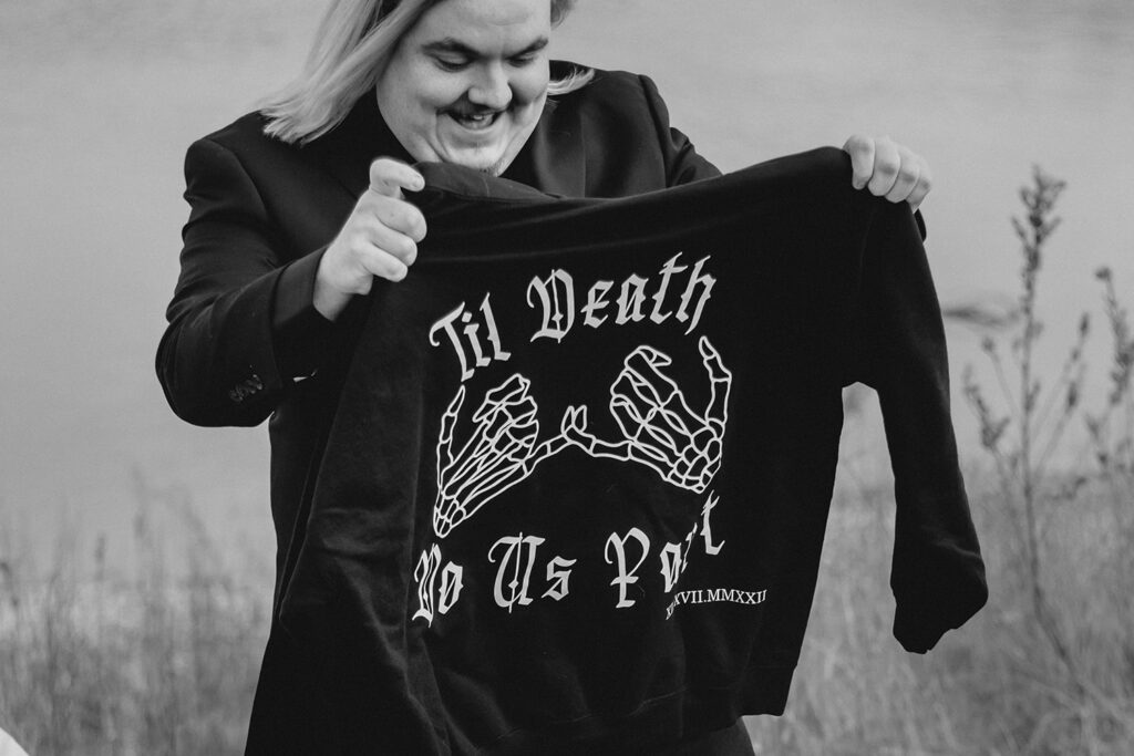groom smiling as he holds up a black hoodies with "Til Death Do Us Part" and an illustration of two skeleton hands with pinkies intertwined
