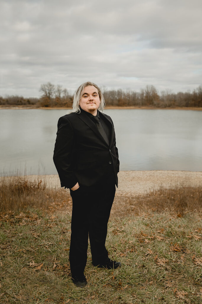 a young groom with shoulder length silver dyed hair and all-black suit smirks at the camera in front of a winter lake