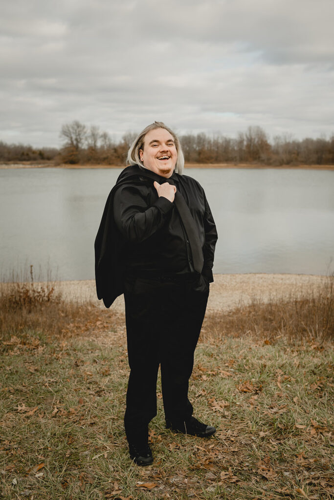 a young groom with shoulder length silver dyed hair and all-black suit laughs at the camera with jacket tossed over his shoulder in front of a winter lake